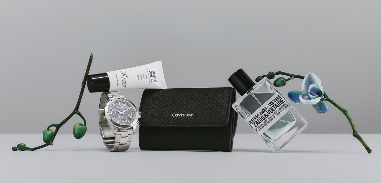 Silverfärgad rostfritt stål klocka, svart läderfodral med Calvin Klein-logotyp, klar parfymflaska och vit tub med ögonkräm med svart lock, på grå bakgrund.