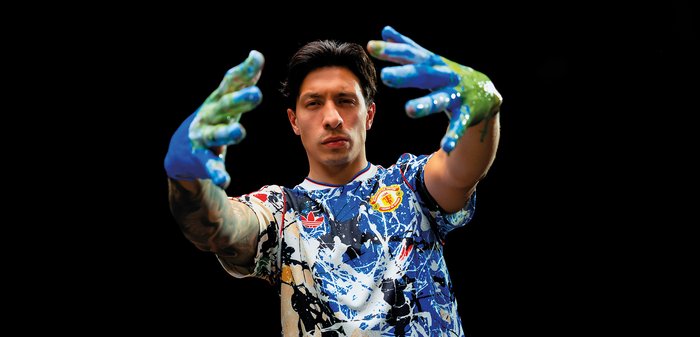 Jeune homme portant une chemise éclaboussée de couleurs, tendant ses mains couvertes de peinture vers le spectateur sur un fond noir.