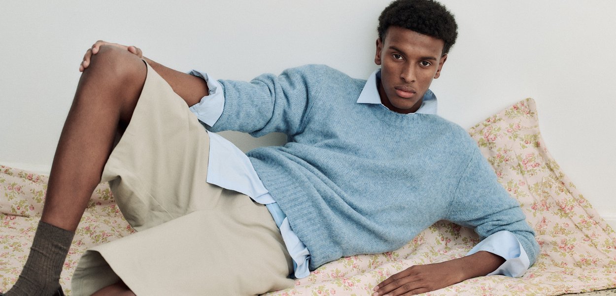 Nuori mies sinisessä neuleessa ja beigeissä shortseissa nojaten kukkakuvioista kangasta vasten valkoista seinää.