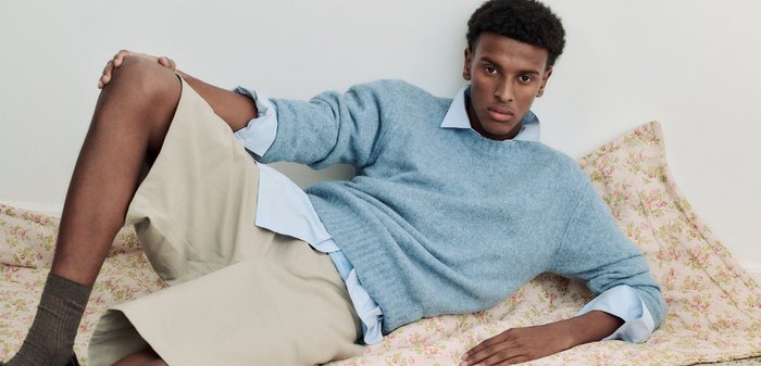 Ung mann i blå genser og beige shorts som lener seg tilbake på blomstrete stoff mot hvit vegg.