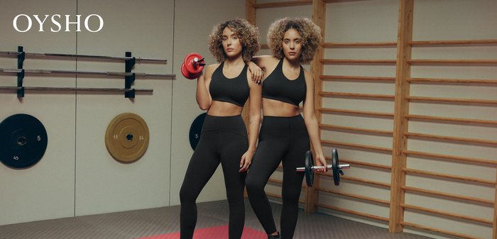 Zwei Frauen in schwarzen Trainingsoutfits halten Gewichte in einem Fitnessstudio, mit Trainingsgeräten und Holzleitern an der Wand im Hintergrund.