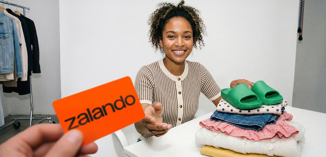 Kobieta uśmiecha się przy stole, sięgając po pomarańczową kartę Zalando, obok niej ułożone są złożone ubrania i zielone sandały.