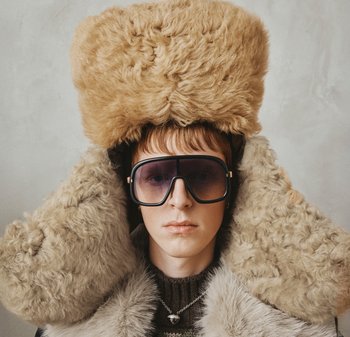 Καπέλο από ψεύτικη γούνα σε καφέ ανοιχτό χρώμα, υπερμεγέθες με υφασμένα διακοσμητικά στοιχεία. Μεγάλα σκουρόχρωμα γυαλιά ηλίου με ορθογώνιο πλαίσιο. Φορεμένα πάνω από έναν γκρι γούνινο γιακά.