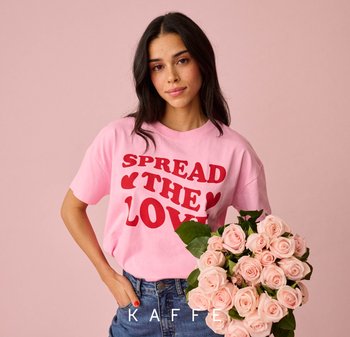 Frau mit dunklem Haar hält einen Strauß rosa Rosen und trägt ein rosa Hemd mit rotem Text "SPREAD THE LOVE" vor hellrosa Hintergrund.