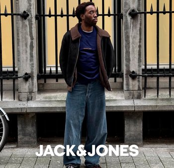 Ein Mann, der eine schwarze Jacke mit braunem Kragen, einen marineblauen Pullover und lockere blaue Jeans trägt, steht vor einer Steinmauer mit schwarzem Eisenzaun.