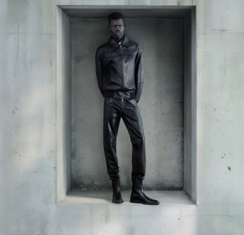 Veste en cuir noir avec col et fermeture éclair devant, associée à un pantalon en cuir noir et des bottines, exposée dans une alcôve en béton.