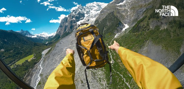 Gelbes Rucksack mit schwarzen Riemen und Beschlägen, gehalten über einer gebirgigen Landschaft mit zerklüfteten Gipfeln, Grünflächen und einem Fluss darunter.