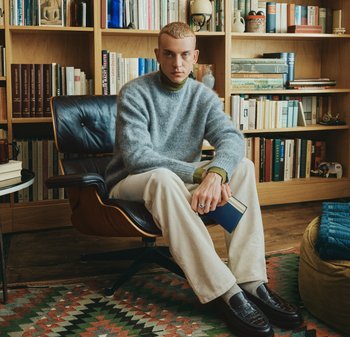 Γκρι μάλλινο πουλόβερ πάνω από πράσινο ζιβάγκο, εκρού παντελόνι από κολόμερο, σκούρα μοκασίνια. Καθισμένος σε μαύρη καρέκλα σε δωμάτιο με βιβλιοθήκες και μοτίβο χαλί.