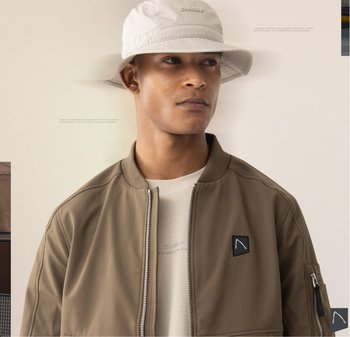 Bruine bomberjack met ritssluiting aan de voorkant, geribbelde kraag en een zwart logo patch. Een lichtgrijze bucket hat maakt de outfit compleet.