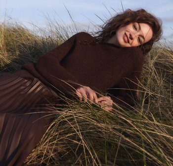 Dunkelbrauner, gestrickter Pullover mit lockerer Passform. Faltenrock in Braun. Liegt inmitten eines grasbewachsenen Geländes vor einem klaren Himmel. Natürliche Texturen.