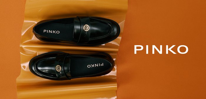 Par de mocasines negros de cuero tipo slip-on con detalle de logo circular dorado, colocados sobre una superficie naranja brillante con el nombre de la marca Pinko visible en el interior de los zapatos.