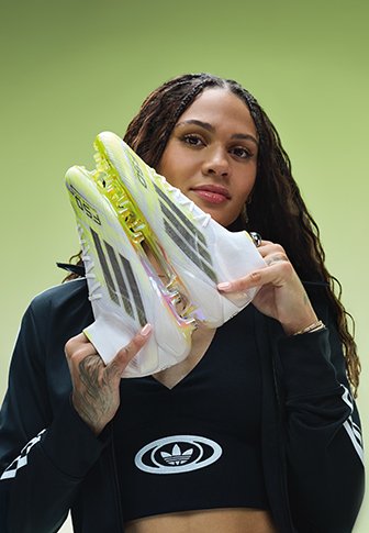 Woman holding white and neon yellow Adidas football boots near her face, wearing black sportswear with Adidas logo on green background.
