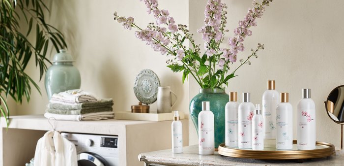 Bouteilles en verre clair avec des motifs floraux, bouchons dorés et argentés sur une table en bois. Fleurs violettes dans un vase vert, machine à laver en arrière-plan.