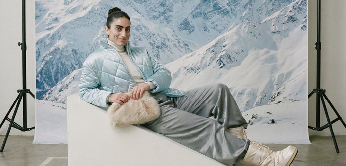 Jeune personne en veste bleu clair et pantalon gris, allongée sur un bloc blanc, tenant un sac en fourrure beige, avec un décor de montagne enneigée dans un studio.