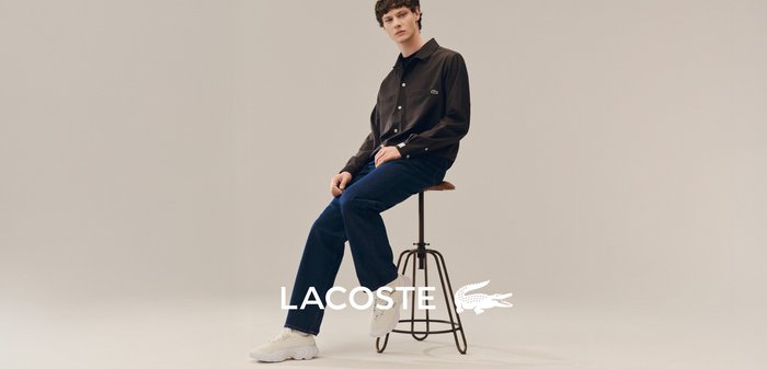 Fiatal férfi ül egy zsámolyon, fekete Lacoste pólót, sötétkék farmert és fehér sportcipőt viselve, egyszínű háttér előtt.
