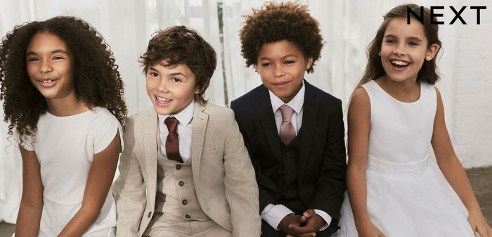 Vier Kinder sitzen, gekleidet in formelle Kleidung. Zwei Mädchen in weißen Kleidern, eines mit Spitzenverzierung. Zwei Jungen in Anzügen: einer beige, einer schwarz mit Weste.