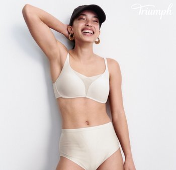 Lächelnde Frau, die einen beigen BH und hoch taillierte Slips, eine schwarze Kappe und goldene Creolen trägt, vor einem weißen Hintergrund posierend.