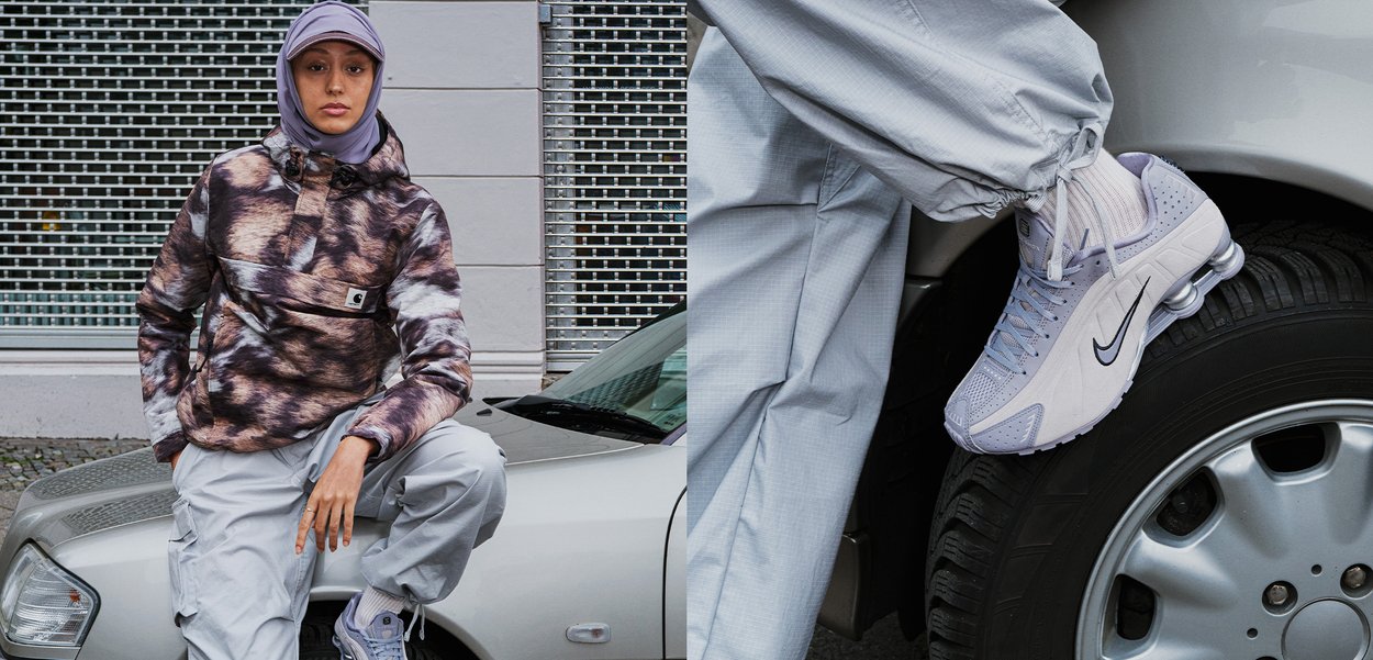 Sneakers Nike de culoare violet deschis cu un swoosh negru, asortați cu pantaloni gri lejerați și o jachetă maro cu model. Fundalul prezintă o mașină.