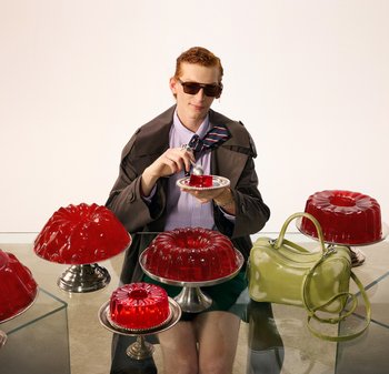 Person som bär solglasögon och trenchcoat äter röd gelédessert vid ett glasbord med flera stora geléformar och en grön handväska.