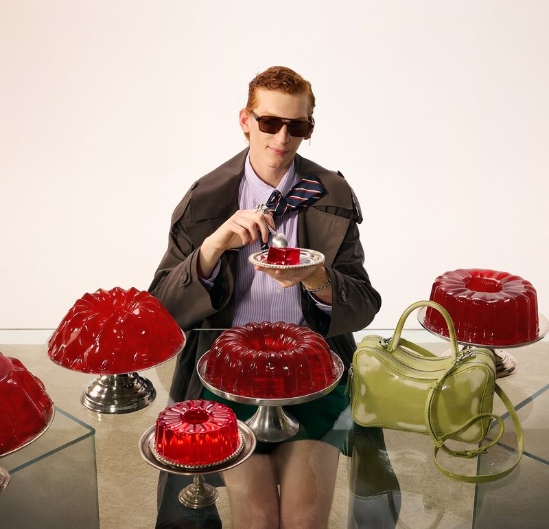 Persona con gafas de sol y chaqueta de traje comiendo un postre de gelatina roja, rodeada de varios moldes de gelatina roja sobre una mesa de cristal.