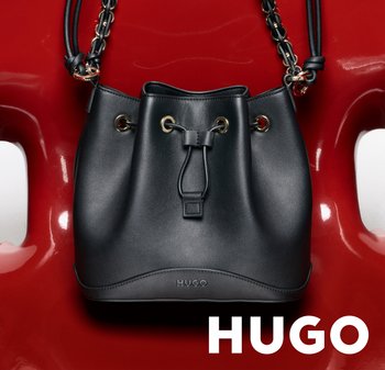Bolso tipo saco de cuero negro con ojales dorados y correa de cadena, con el logo "HUGO" en relieve, exhibido sobre una silla roja brillante.