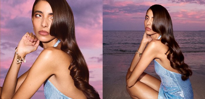 Femme aux longs cheveux foncés en robe bleue posant sur une plage au coucher du soleil avec un ciel rose et violet reflété dans l'eau.