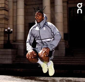Sportler mit Dreadlocks springt vor Steinsäulen, trägt eine graue Jacke, schwarze Shorts, weiße Socken und neongelbe Laufschuhe.
