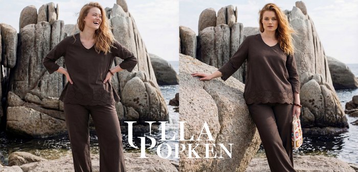 Woman with wavy blonde hair wearing dark brown top and pants stands against large coastal rocks, holding a colorful crochet handbag by the sea.