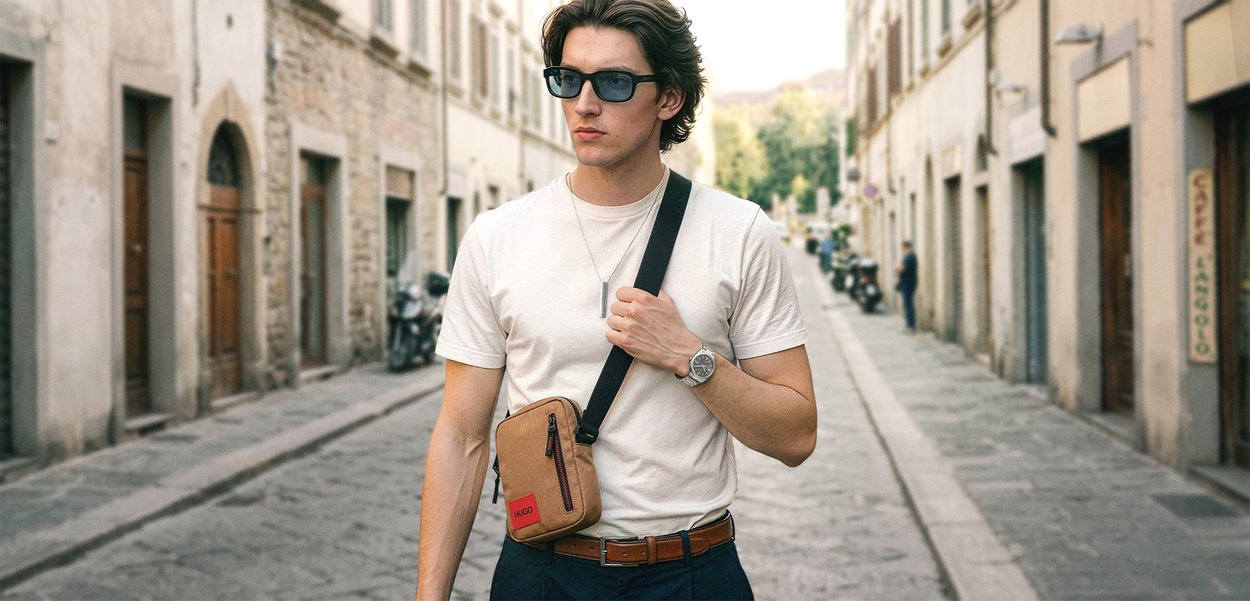 Hombre con gafas de sol, camiseta blanca y bolso cruzado caminando por una calle adoquinada bordeada de edificios históricos.