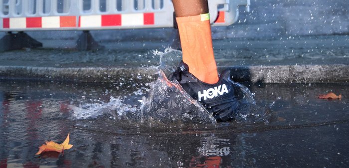 A black HOKA running shoe splashing in water, adorned with a bright orange sock. The wet ground mirrors droplets and features a nearby fallen leaf.