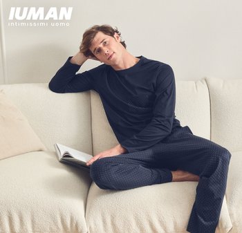 Uomo in pigiama blu navy a pois sdraiato su un divano crema, che tiene un libro aperto, con testo "Iuman Intimissimi Uomo" in alto a sinistra.