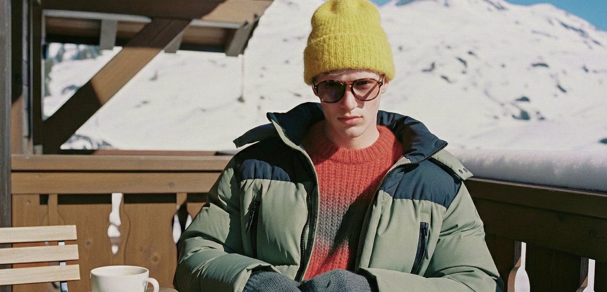 Πράσινο και ναυτικό μπουφάν με φερμουάρ στις τσέπες, πορτοκαλί πουλόβερ με διαβάθμιση, γκρι γάντια, κίτρινο σκουφάκι και γυαλιά ηλίου. Χιονισμένο εξωτερικό περιβάλλον.