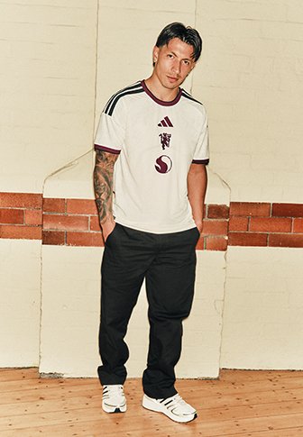 Mann mit tätowiertem Arm, der ein weißes Sporttrikot, schwarze Hose und weiße Sneakers trägt und drinnen vor einer beigen und ziegelsteinernen Wand steht.