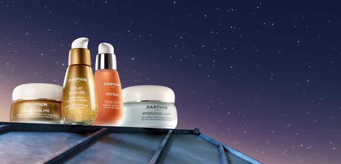 Quatre produits de soins de la peau de Darphin : un sérum doré, un sérum orange et deux pots de crème (blanc et argent, avec des accents dorés), présentés sur un fond étoilé.