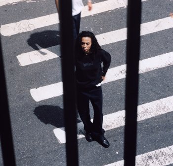 Persona con cabello rizado vistiendo ropa negra, de pie en un paso de peatones, visto a través de barras verticales negras que proyectan sombras en la acera.