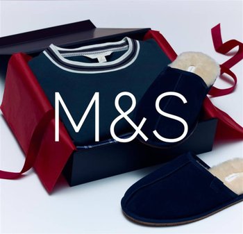 Granatowe kapcie z futrzanym wykończeniem obok ciemnozielonego swetra z białym paskiem przy dekolcie, zapakowane w pudełko prezentowe z czerwonymi akcentami.