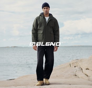 Giacca con cappuccio verde oliva con due tasche frontali, pantaloni neri e scarpe beige, in piedi su un terreno roccioso vicino all'acqua.