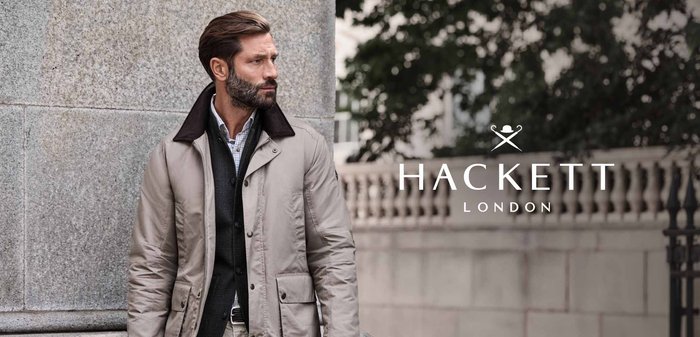 Veste beige clair avec un col marron, portée sur un pull sombre et une chemise à carreaux. Dispose de poches et d'une fermeture éclair devant un mur en pierre.