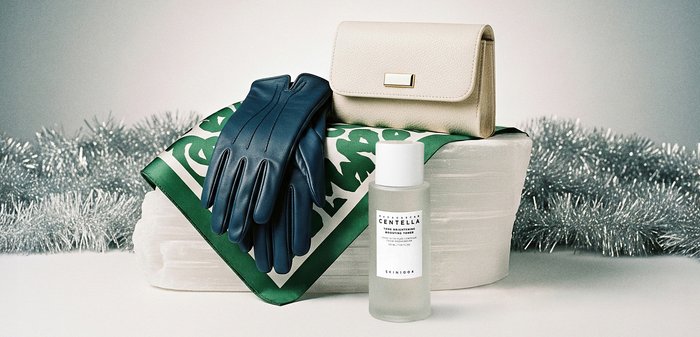 Blå läderhandskar, krämfärgad clutchtaske, grönmönstrad sidenhalsduk och en genomskinlig tonerkanna arrangerade på en strukturerad vit yta.