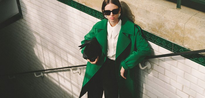 Femme portant un manteau vert, une chemise blanche, un pantalon noir et des lunettes de soleil, se tenant sur des escaliers dans un métro carrelé, tenant un sac noir.