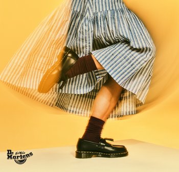 Mocasines de cuero negro con acabado brillante, que presentan un fleco decorativo y costuras amarillas, combinados con calcetines burdeos con canalé y una falda a rayas.