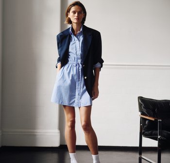 Vrouw staat binnen, draagt een blauw-wit gestreepte jurk met een marineblauwe blazer en witte sokken, naast een zwarte leren stoel.