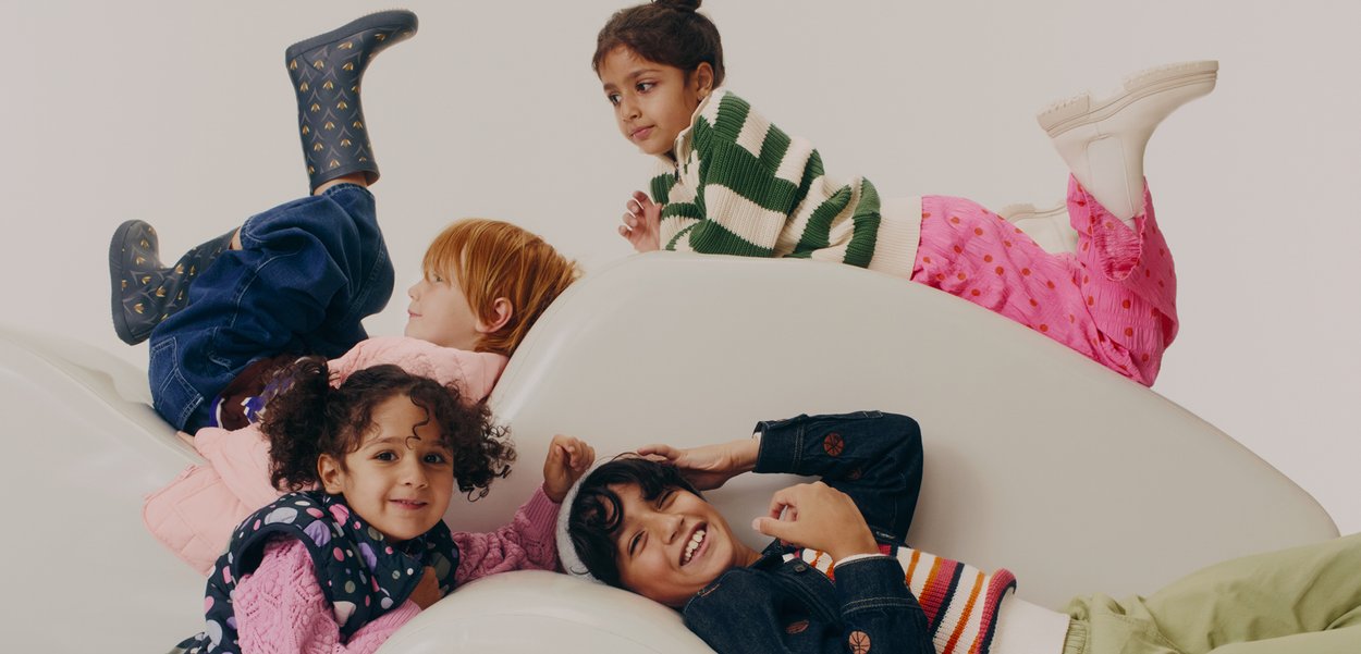 Patru copii stau întinși pe o suprafață curbată, albă și netedă. Aceștia poartă haine colorate, inclusiv un pulover verde cu dungi și pantaloni cu modele.