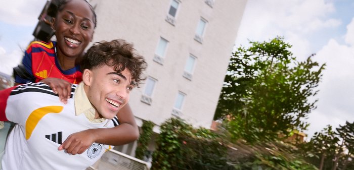 Νέος άνδρας δίνει καβάλα σε μια χαμογελαστή νεαρή γυναίκα σε εξωτερικό χώρο κοντά σε κτήριο και φυλλώματα κάτω από έναν μερικώς συννεφιασμένο ουρανό.