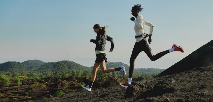 Runner wearing white long-sleeve shirt and red shoes jogging on rocky, sparse vegetation trail in outdoor natural setting.