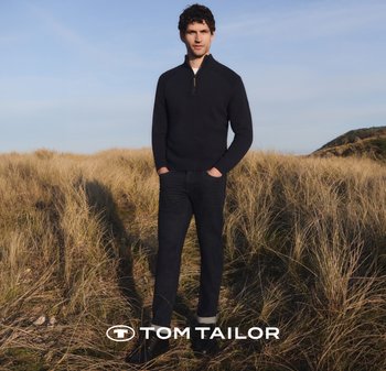 Granatowy sweter zapinany na zamek z fakturowanym splotem, ciemne dżinsy i czarne buty. Stojący na wysokiej trawie, na tle niebieskiego nieba.