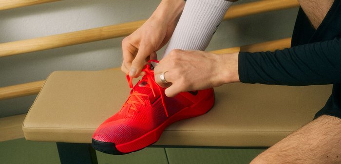 Személy élénkpiros sportcipőt kötöz, miközben egy bézs padon ül, fehér zoknit és fekete ujjú felsőt visel.