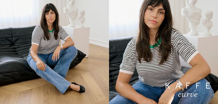 Mladá žena s dlhými tmavými vlasmi v pruhovanom tričku a džínsoch sediaca na čiernej koženej pohovke s bielymi sochami v pozadí.