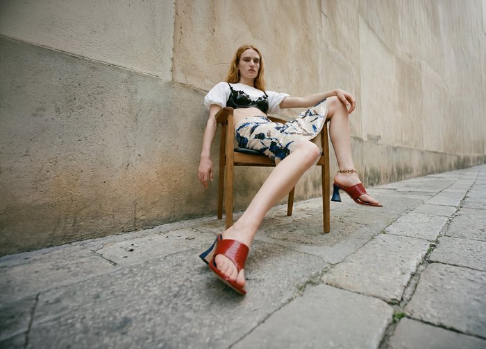 Femme aux cheveux roux assise sur une chaise en bois sur un pavement en pierre, vêtue d'un haut blanc, d'une jupe fleurie, d'un soutien-gorge noir et de sandales à talons rouges.