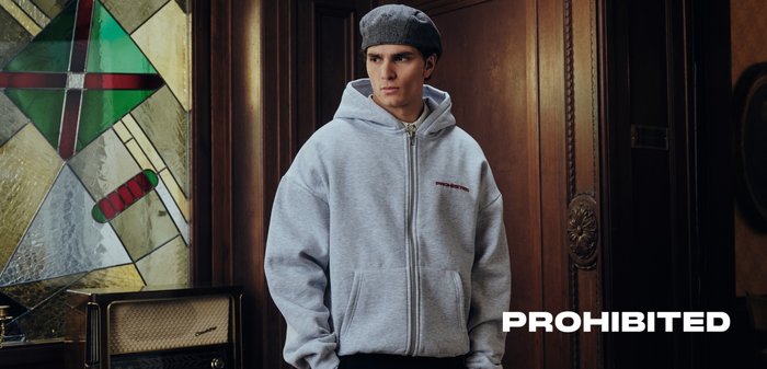Young man wearing grey zip-up hoodie and beret stands indoors near stained glass window and vintage radio, with "PROHIBITED" text.